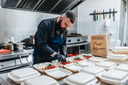 Un chef remplit plusieurs barquettes à emporter posées sur le plan de travail de la cuisine d'un restaurant ; un sac portant l'inscription « livraison de repas » est posé à côté. 